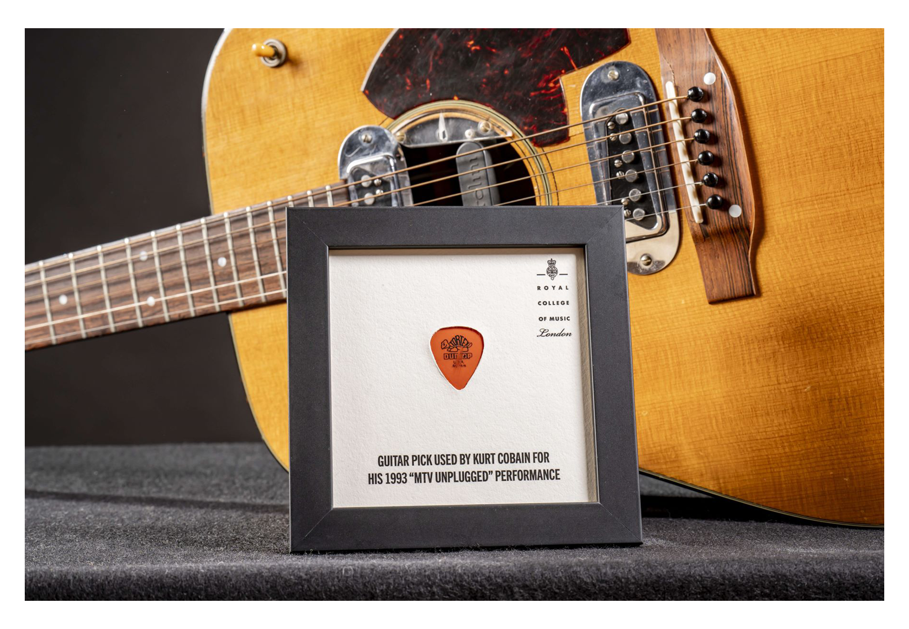 A plectrum in a frame, with a classic guitar in the background, with grey background.